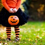 child in orange shirt, black skirt, and orange and black stripe stockings holding a jack-o-lantern for trick or treating